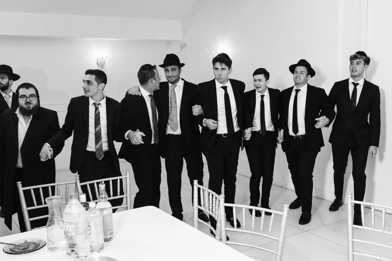 Black-and-white photo of a group of men in formal black suits and striped ties walking/posing together indoors beside a table with bottles. Wedding photography at Riverview Ballroom, Cliffside Park, NJ.