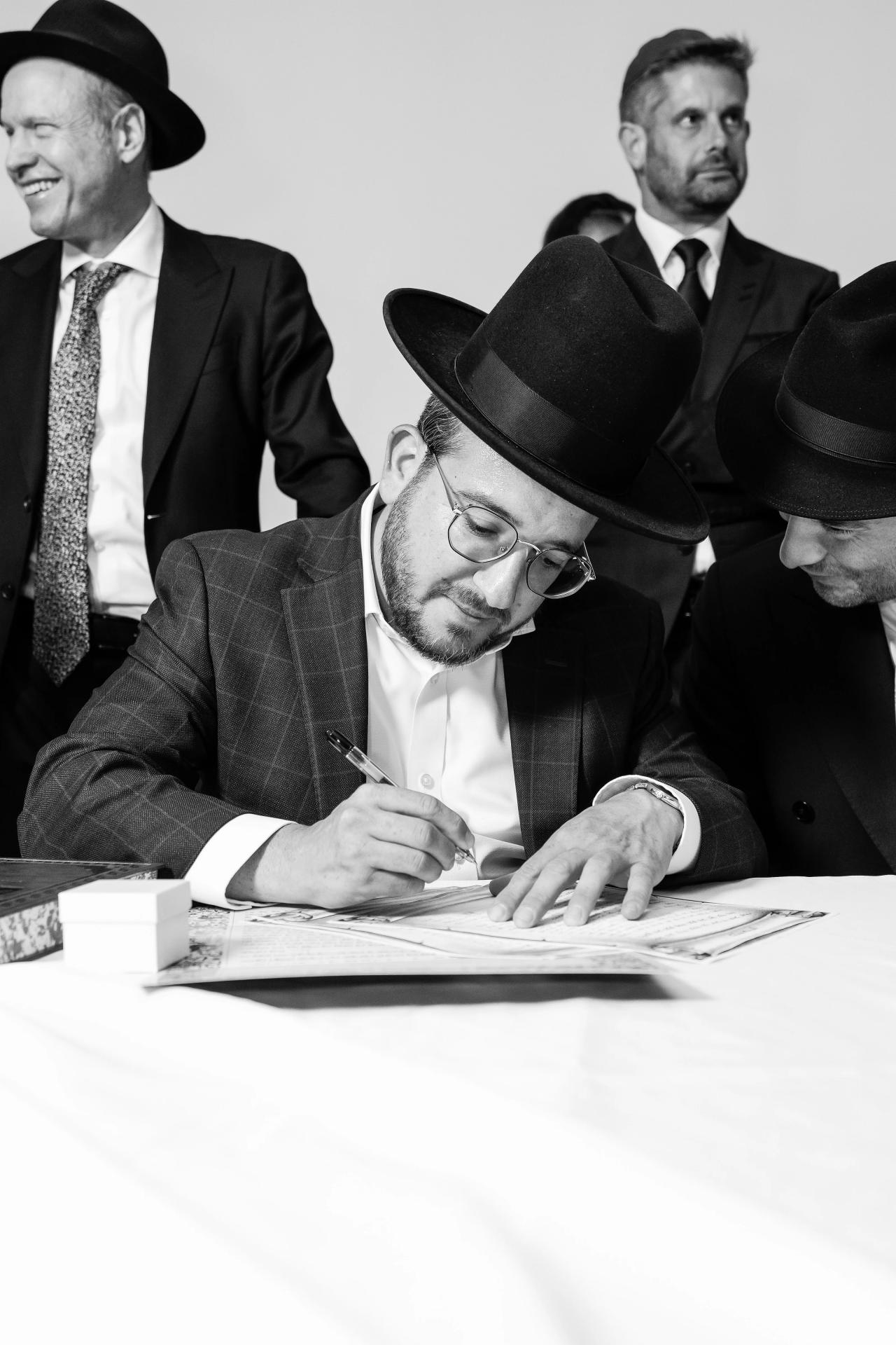 Black-and-white close-up of a man signing the ketubah at a table, with the document and pen in view. Wedding photography at Riverview Ballroom, Cliffside Park, NJ.