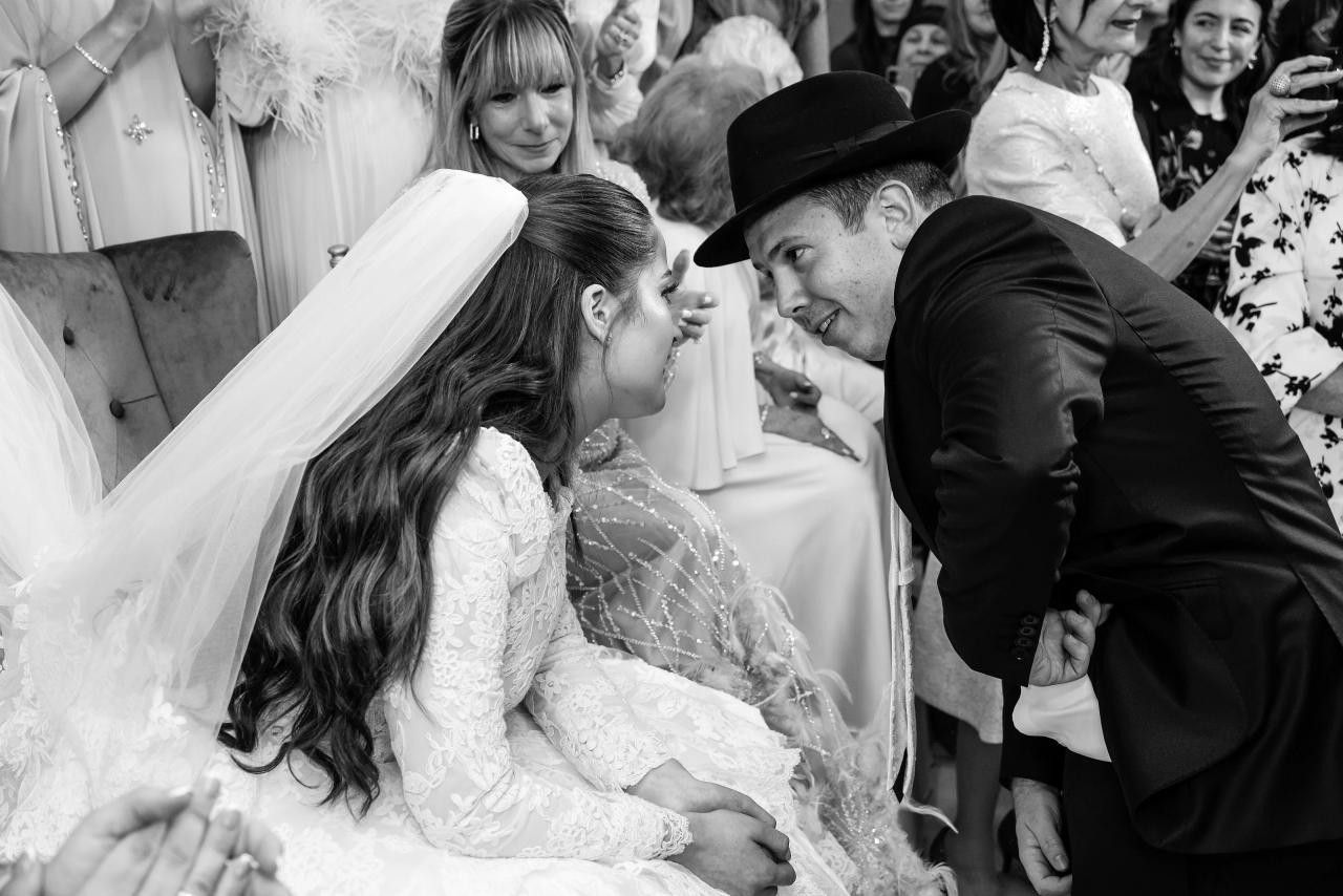 An intimate black-and-white bedeken moment, with the seated bride, a family blessing and guests watching closely behind. Wedding photography at Riverview Ballroom, Cliffside Park, NJ.