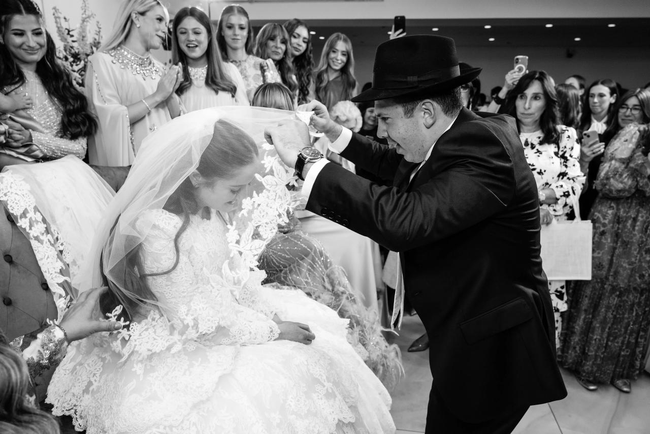 Black-and-white bedeken moment as the veil falls around the seated bride, with family watching behind. Wedding photography at Riverview Ballroom, Cliffside Park, NJ.