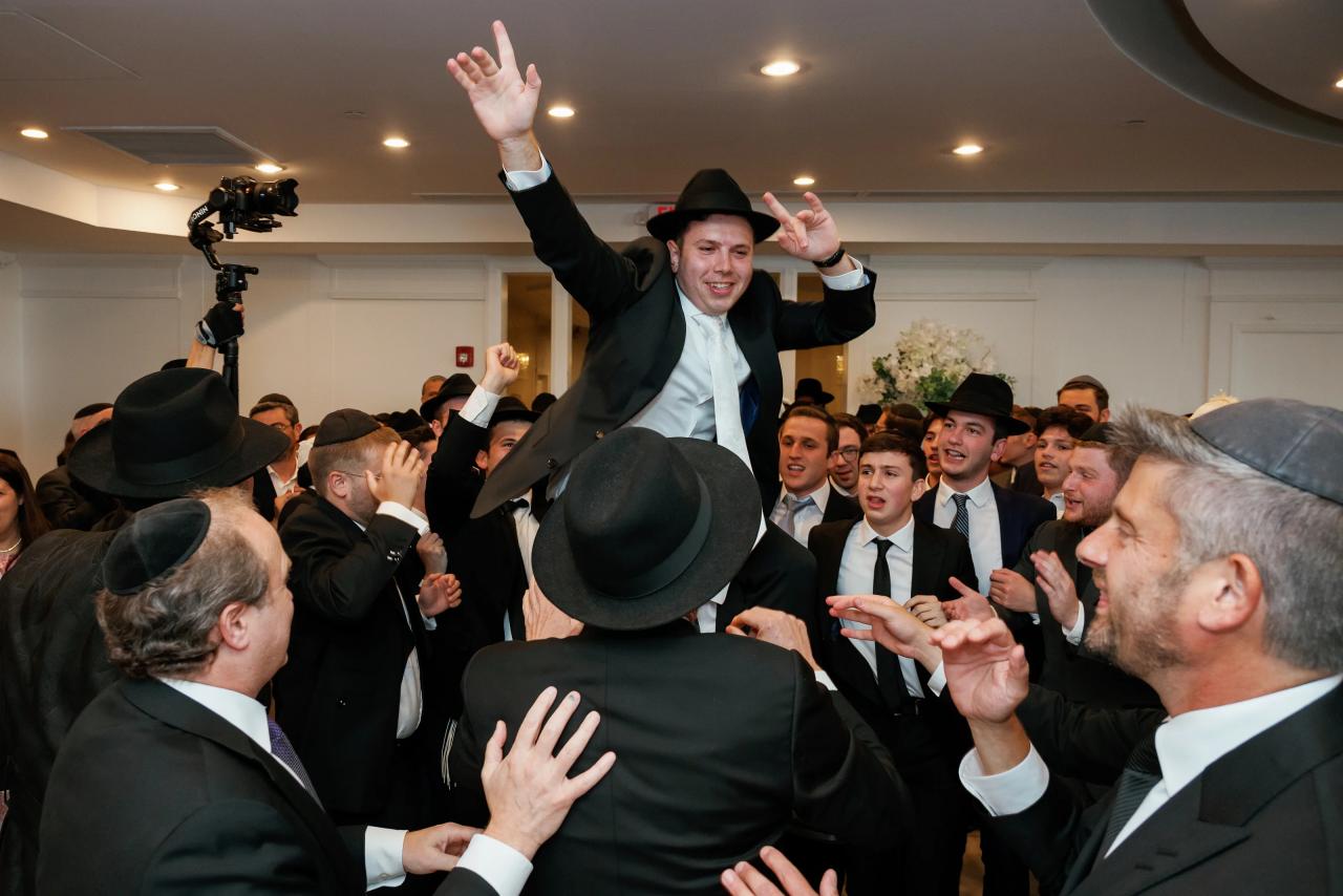 Smiling groom in a hat raises his hand in a crowded indoor celebration as guests cheer and gesture around him. Wedding photography at Riverview Ballroom, Cliffside Park, NJ.