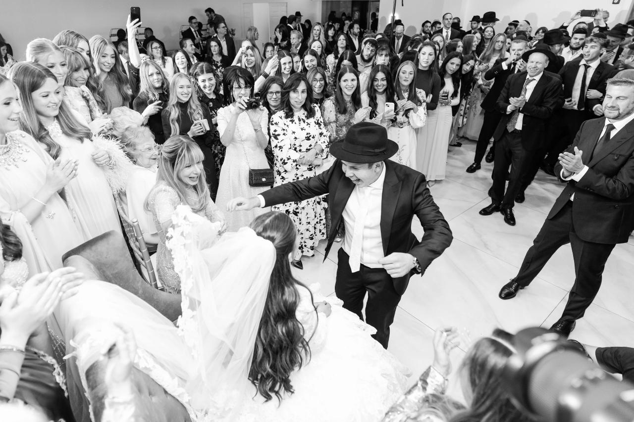 Black-and-white bedeken scene with the seated bride surrounded by smiling, cheering family and friends. Wedding photography at Riverview Ballroom, Cliffside Park, NJ.