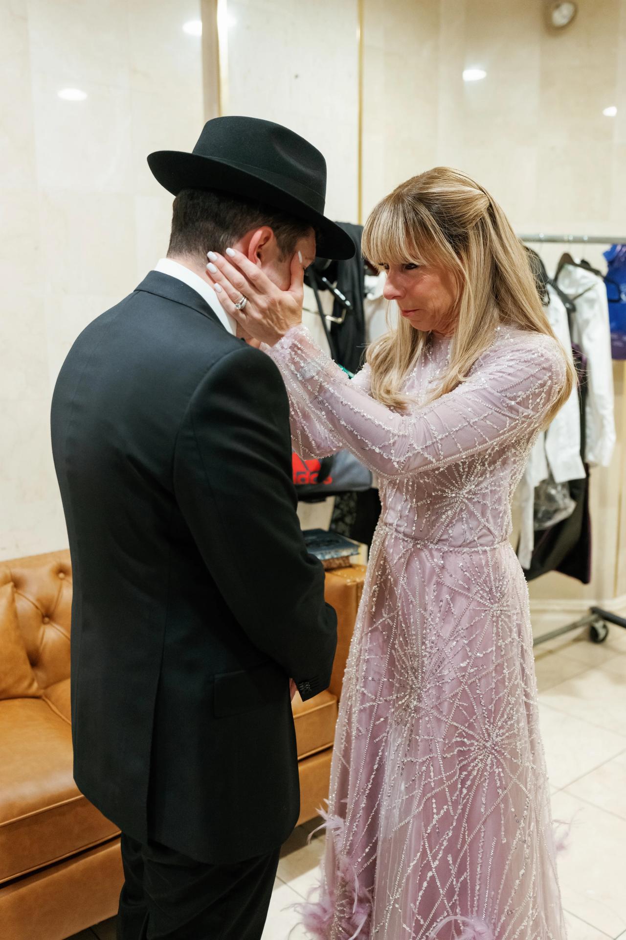 A family member tenderly holds the groom’s face during a quiet pre-ceremony blessing indoors. Wedding photography at Riverview Ballroom, Cliffside Park, NJ.