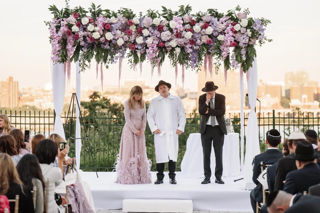 Wedding ceremony under a floral chuppah on an outdoor stage with guests seated and a city skyline in the background. Wedding photography at Riverview Ballroom, Cliffside Park, NJ.
