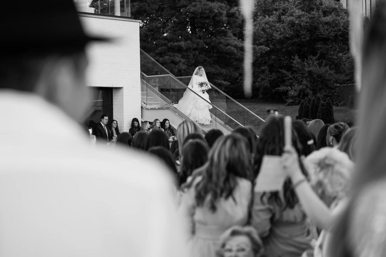 Black-and-white wedding ceremony aisle scene: bride on an elevated walkway holding a bouquet as guests in the foreground watch from a distance. Wedding photography at Riverview Ballroom, Cliffside Park, NJ.
