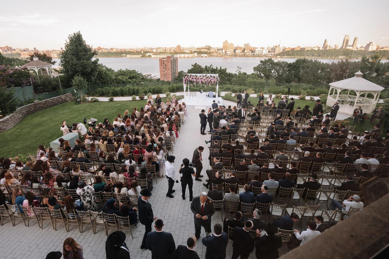 Wide overhead view of a wedding ceremony with a chuppah at the far end and guests seated along a central aisle outdoors. Wedding photography at Riverview Ballroom, Cliffside Park, NJ.