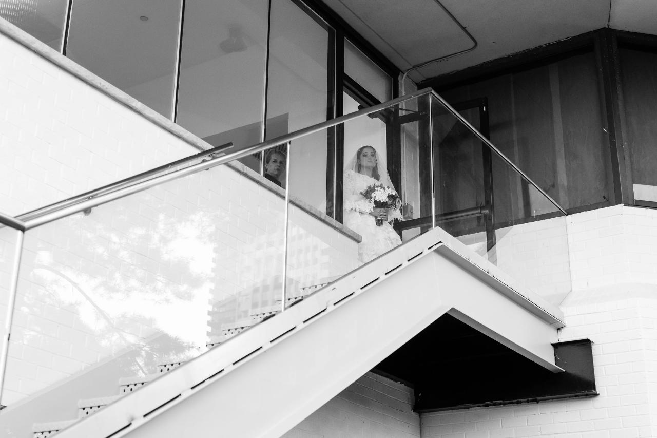 Black-and-white architectural stairwell view with a bride visible through the upper window, framed by diagonal railings and bright light below. Wedding photography at Riverview Ballroom, Cliffside Park, NJ.