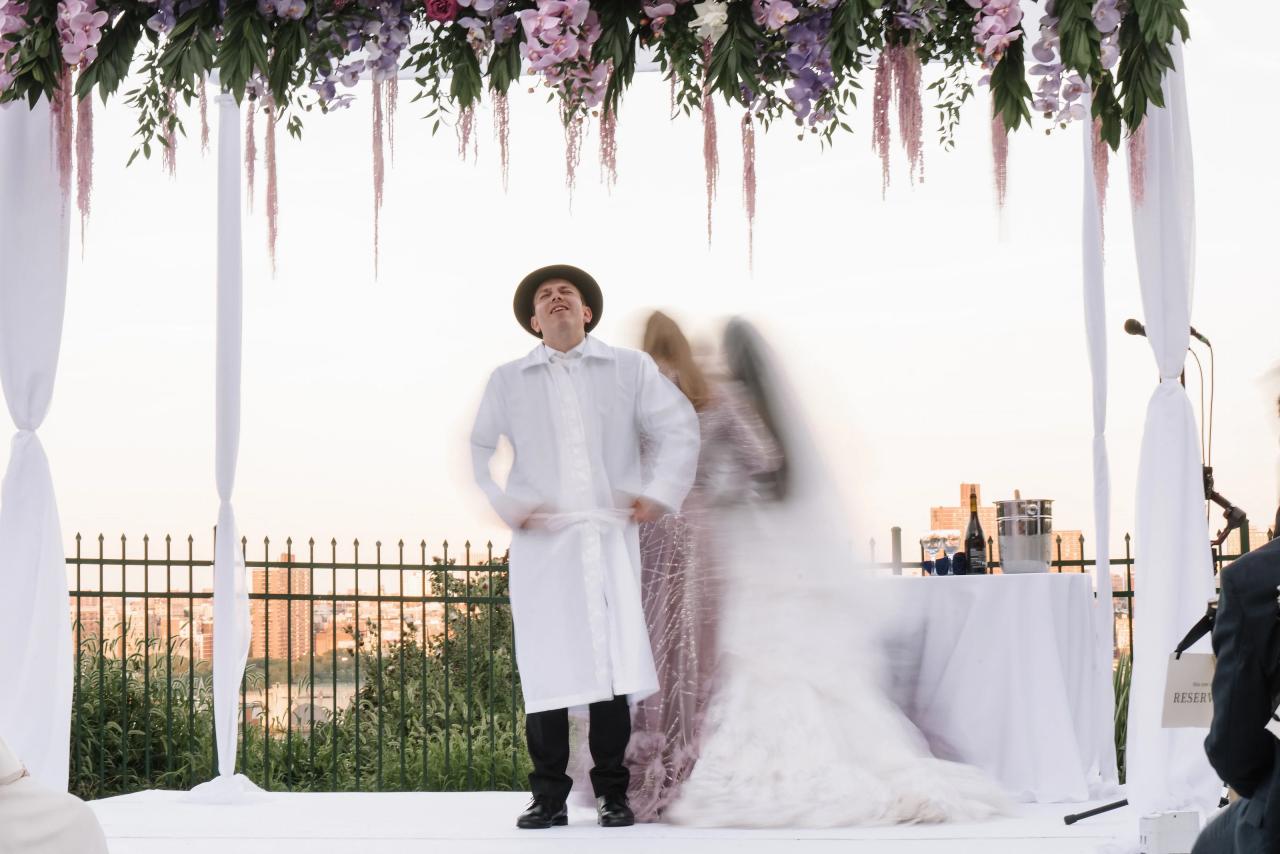 The groom remains relatively still beneath a floral chuppah while the bride’s circling movement is intentionally blurred in warm riverfront light. Wedding photography at Riverview Ballroom, Cliffside Park, NJ.