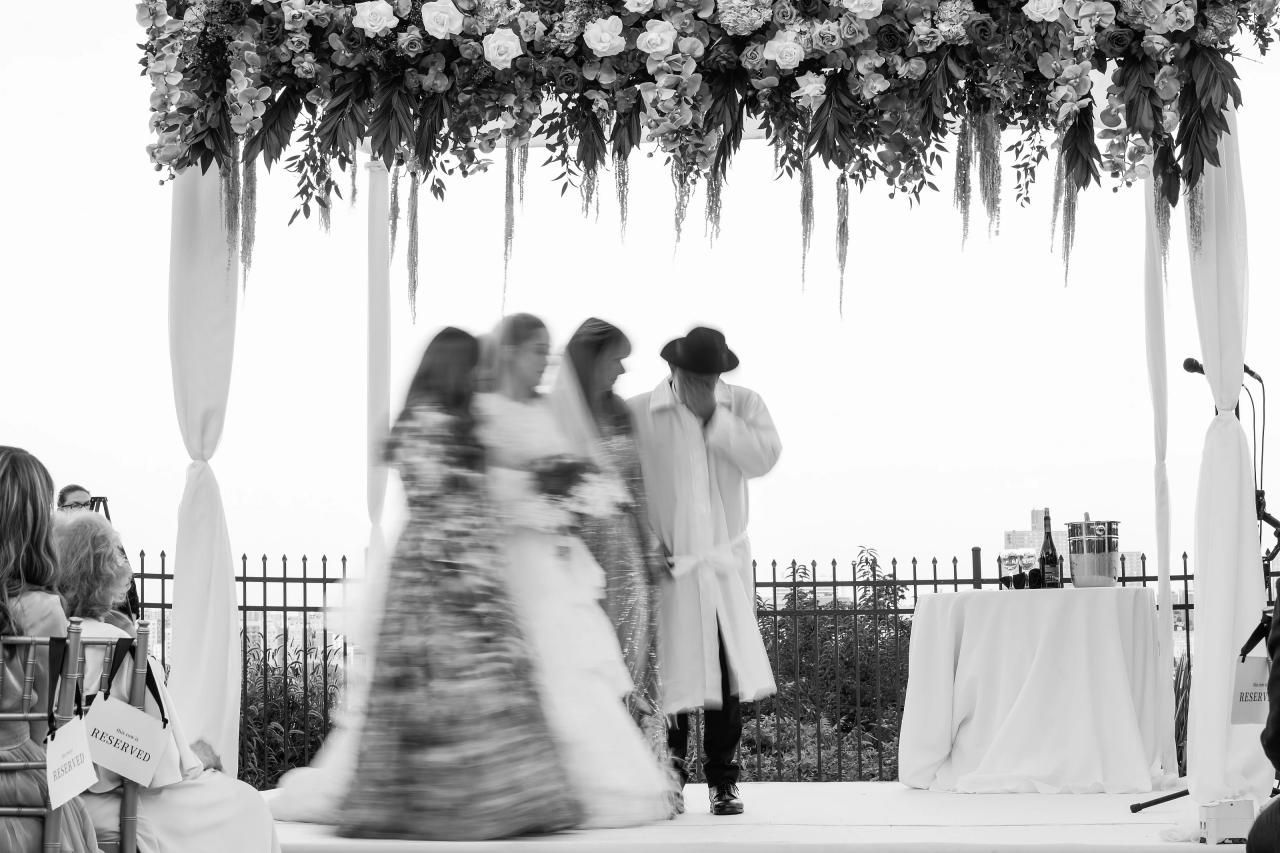 Warm sunset light on the groom beneath a floral chuppah, with the riverfront skyline behind. Wedding photography at Riverview Ballroom, Cliffside Park, NJ.