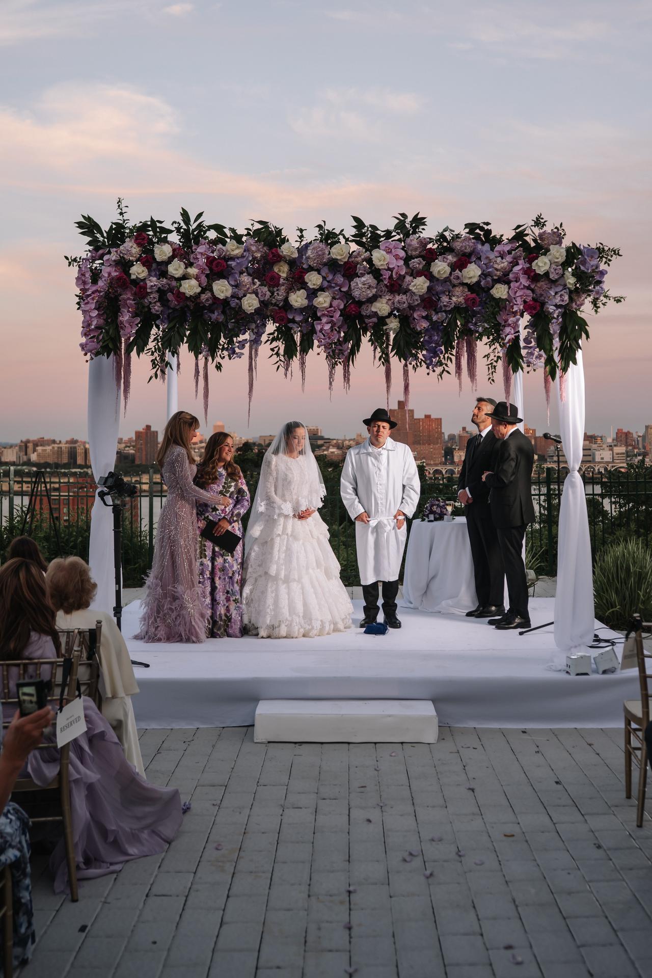 A floral chuppah at dusk on the riverfront terrace, with the skyline fading into lavender and blue. Wedding photography at Riverview Ballroom, Cliffside Park, NJ.