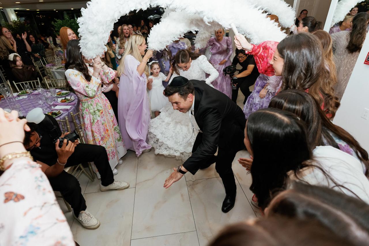 An overhead view of the newlyweds entering beneath a white floral arch, surrounded by celebrating guests. Wedding photography at Riverview Ballroom, Cliffside Park, NJ.