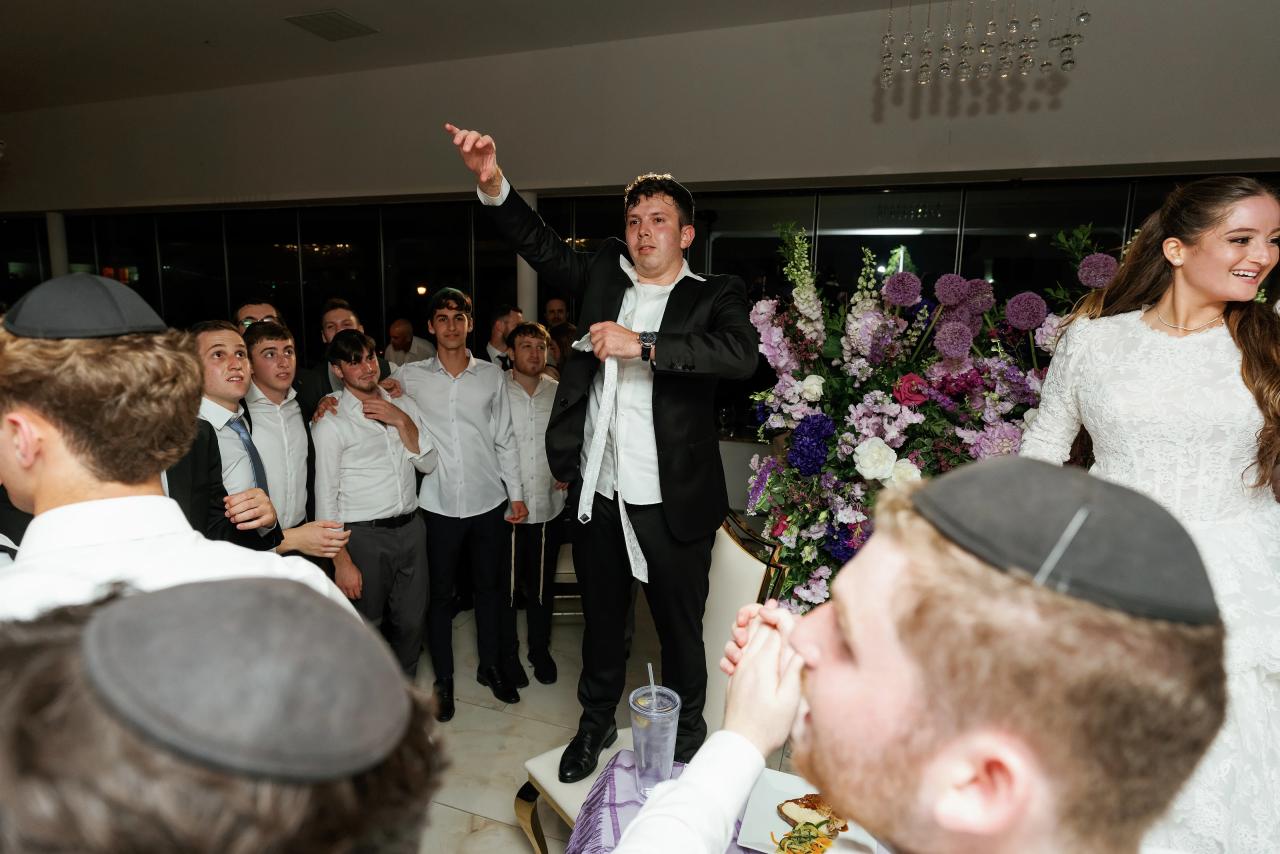 The groom speaks with the bride seated nearby, surrounded by wedding guests and raised hands. Wedding photography at Riverview Ballroom, Cliffside Park, NJ.