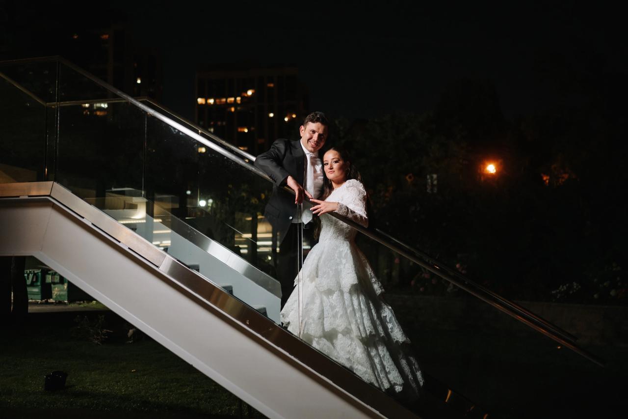 The newlyweds on a nighttime staircase, framed by diagonal glass rails and warm reflections. Wedding photography at Riverview Ballroom, Cliffside Park, NJ.