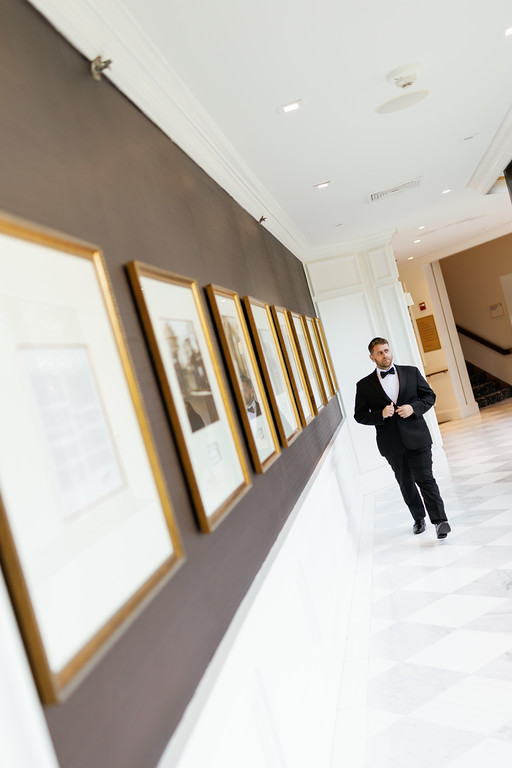 A groom in a black suit adjusts his cuff while walking down a bright corridor with framed pictures on the wall.