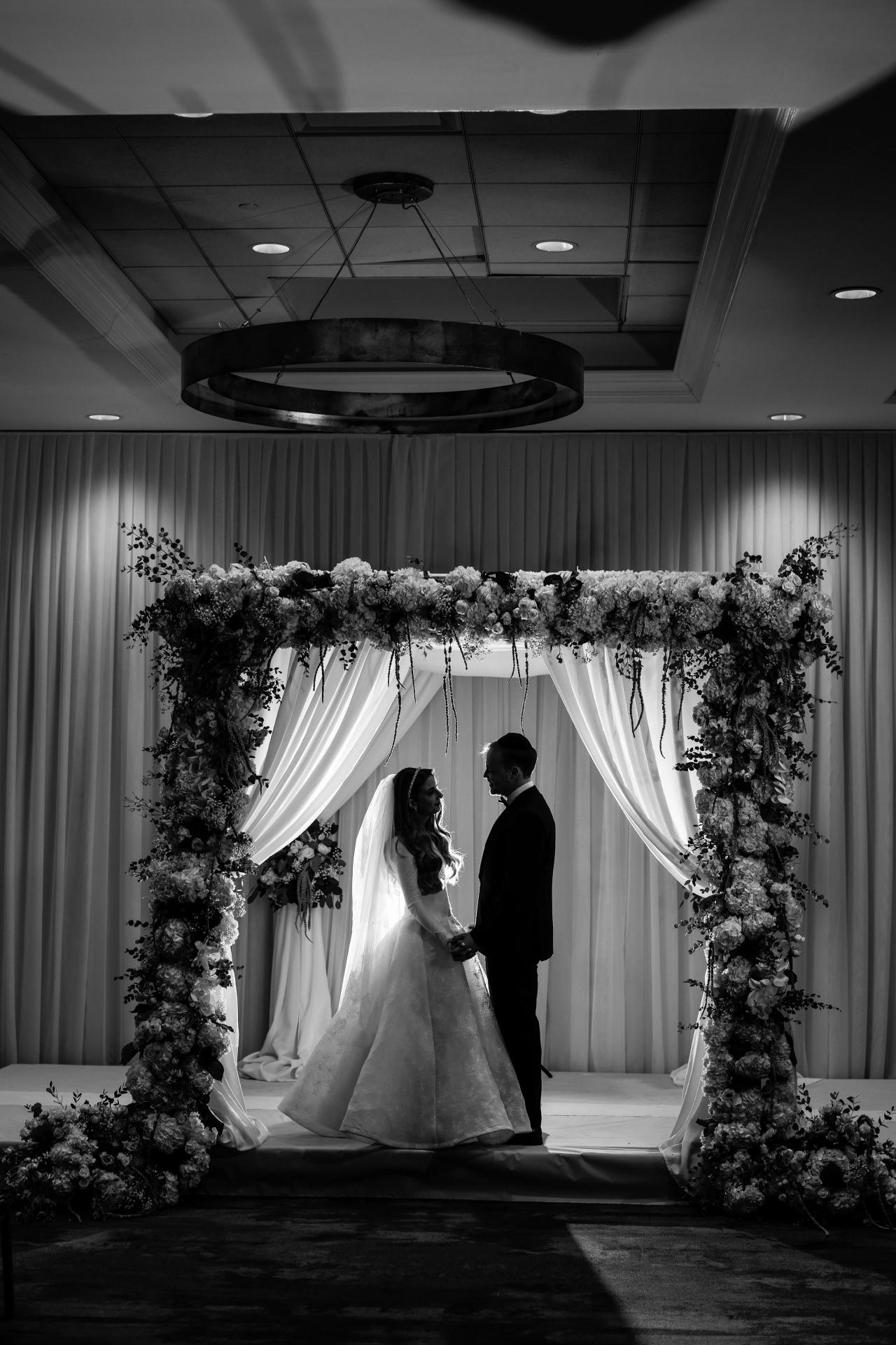 Black-and-white couple portrait beneath a flower-framed chuppah. Wedding photography at Brightstone, Passaic, NJ.