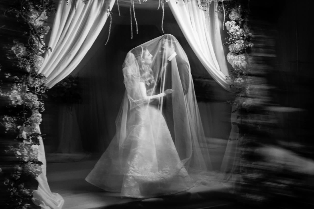 The couple beneath a veil with expressive black-and-white radial movement around the floral arch. Wedding photography at Brightstone, Passaic, NJ.