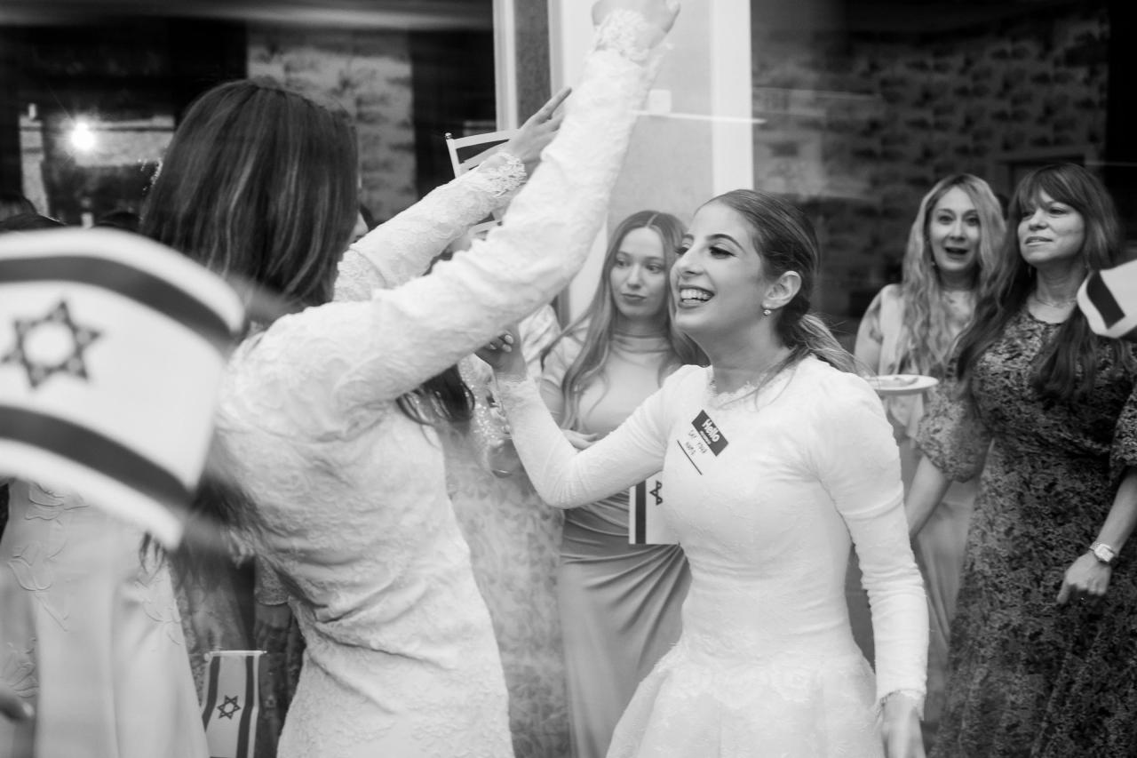 Black-and-white dance portrait with the smiling bride and animated guests in the foreground. Wedding photography at Brightstone, Passaic, NJ.