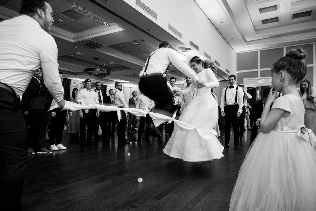 The couple jumps over a rope in a black-and-white dance-floor photograph with a young guest watching. Wedding photography at Brightstone, Passaic, NJ.
