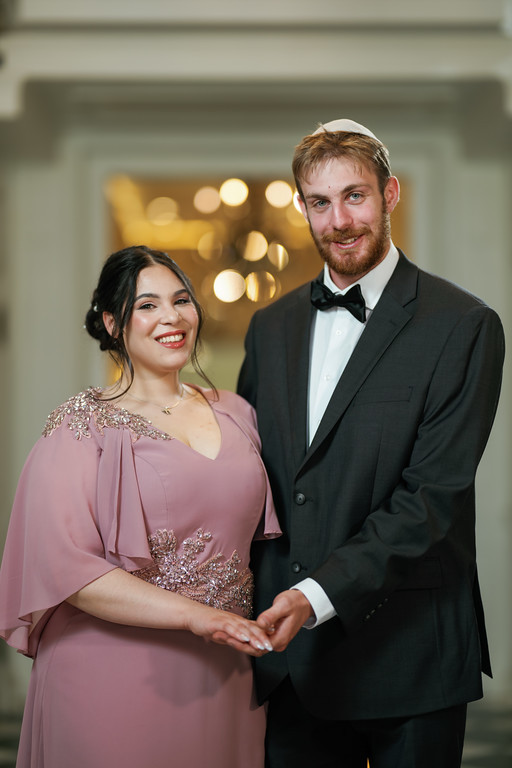 Smiling bride and groom in formal attire holding hands indoors during a wedding event.