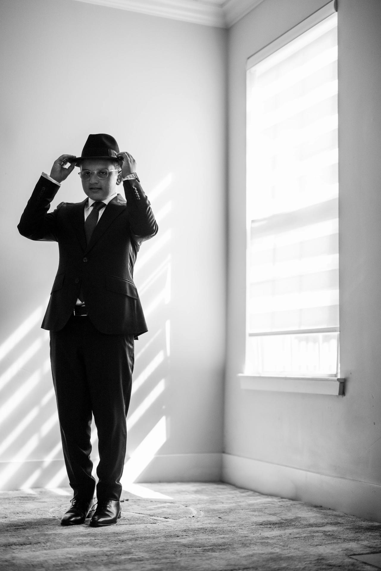 Black-and-white full-length portrait of the bar mitzvah boy adjusting his hat beside a bright window, before the Eagle Ridge bar mitzvah. Bar Mitzvah photography.
