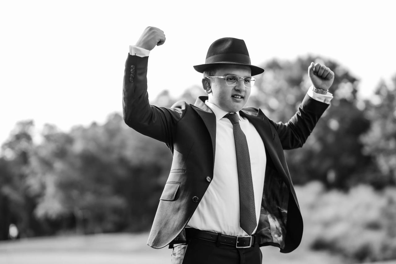 The bar mitzvah boy raises his arms outdoors in a playful black-and-white portrait. Bar Mitzvah photography at Eagle Ridge, Lakewood, NJ.
