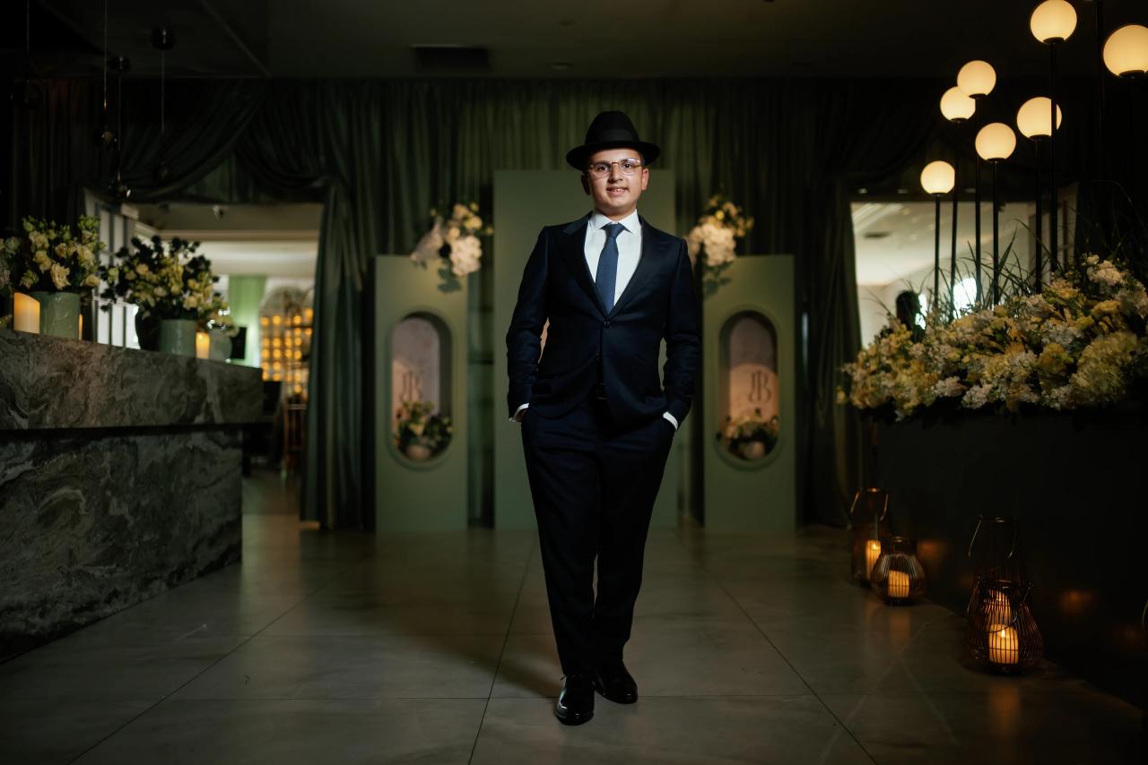 Full-length portrait of the bar mitzvah boy between green drapery, flowers and glowing globe lights. Bar Mitzvah photography at Eagle Ridge, Lakewood, NJ.