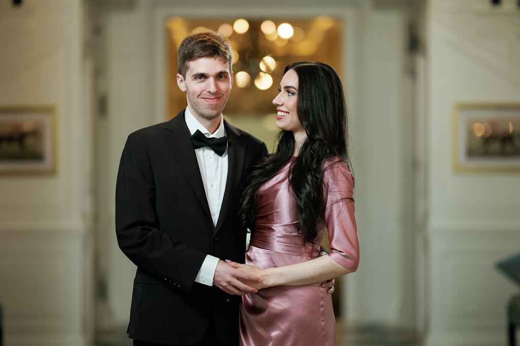 Smiling groom and bride standing together in formal attire, holding hands and looking at each other in a softly lit room.
