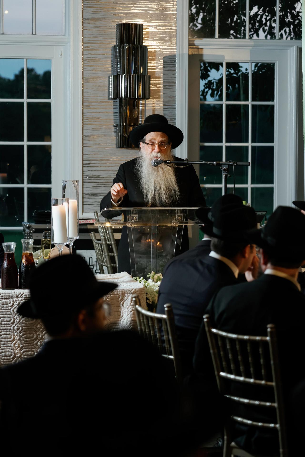 An elder speaks at a microphone, framed by seated guests and tall windows. Bar Mitzvah photography at Eagle Ridge, Lakewood, NJ.