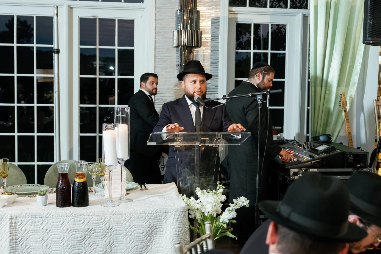 The bar mitzvah boy stands beside the microphone with guests and tall windows behind him. Bar Mitzvah photography at Eagle Ridge, Lakewood, NJ.
