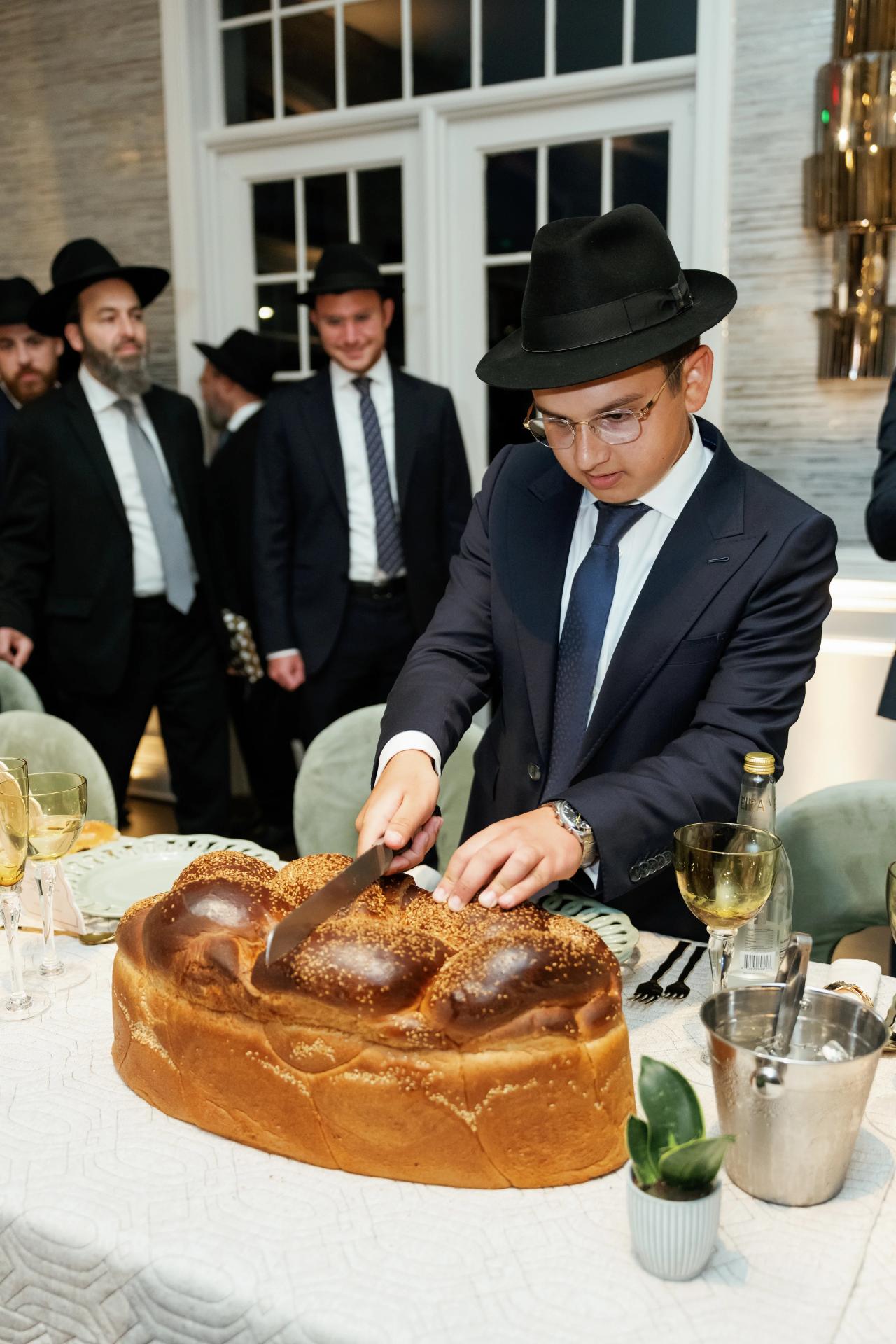 The bar mitzvah boy cuts a large braided challah at the reception table. Bar Mitzvah photography at Eagle Ridge, Lakewood, NJ.