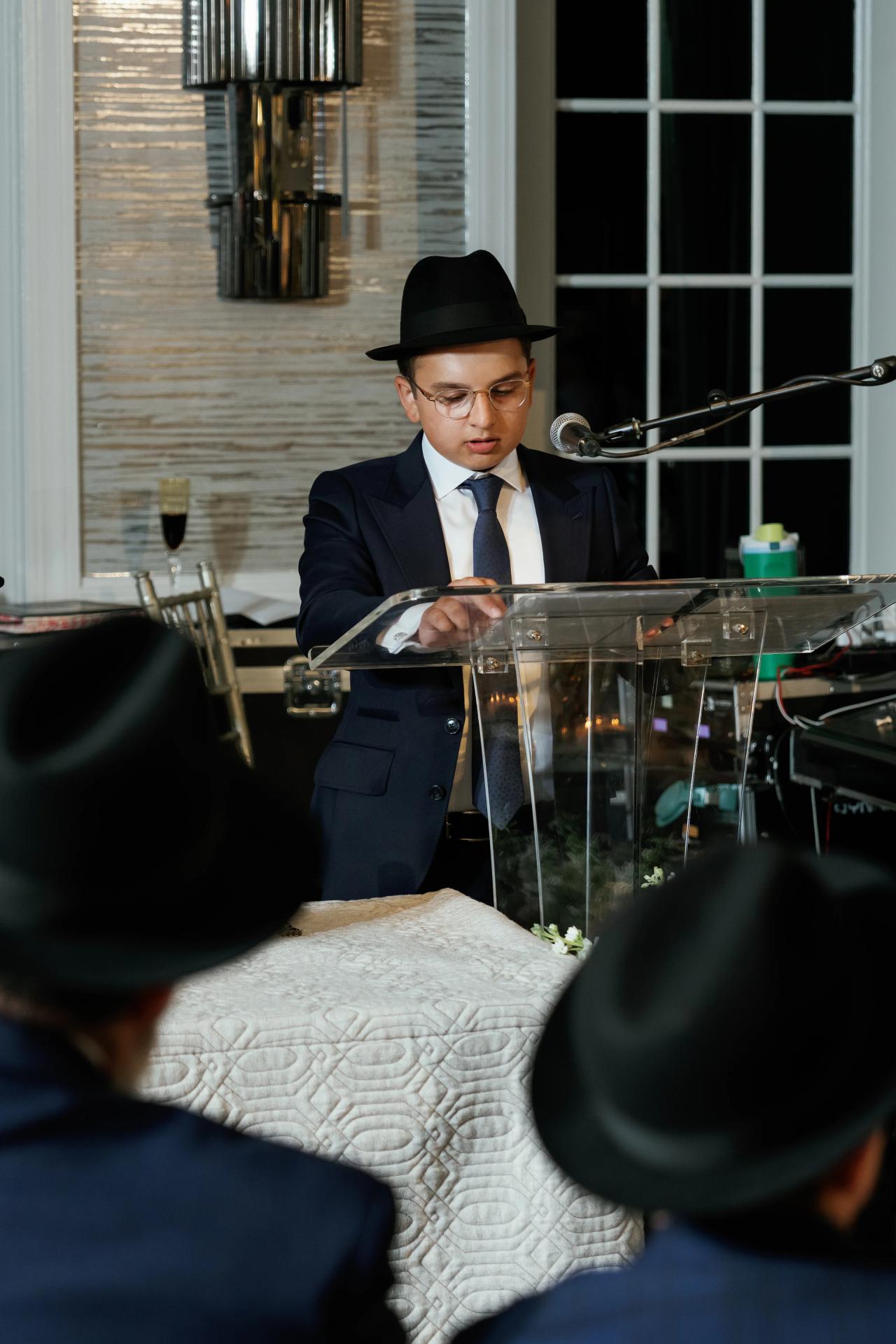 The bar mitzvah boy speaks at a clear lectern, framed by windows and seated guests. Bar Mitzvah photography at Eagle Ridge, Lakewood, NJ.