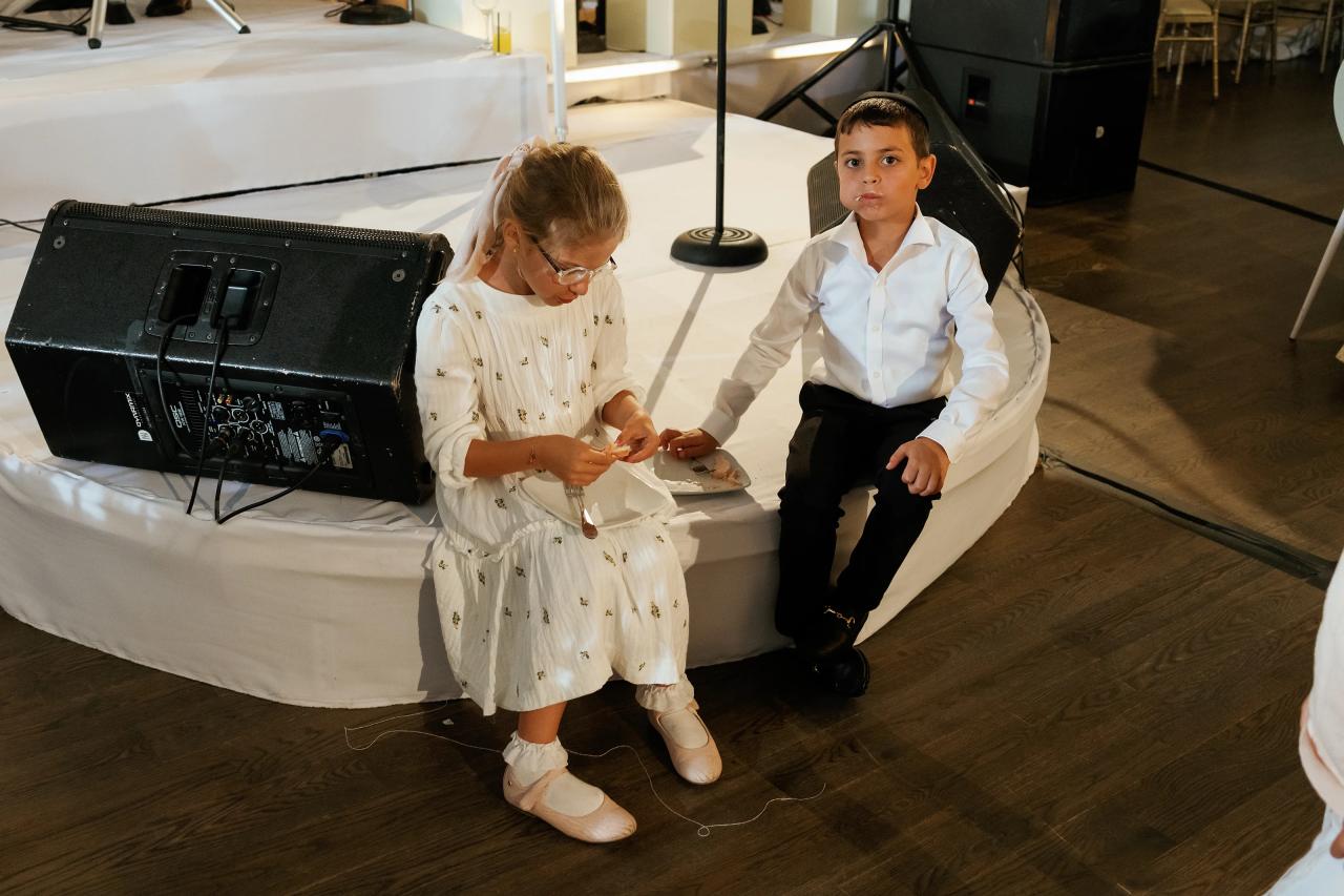 Two children sit together near the band during the bar mitzvah reception. Bar Mitzvah photography at Eagle Ridge, Lakewood, NJ.