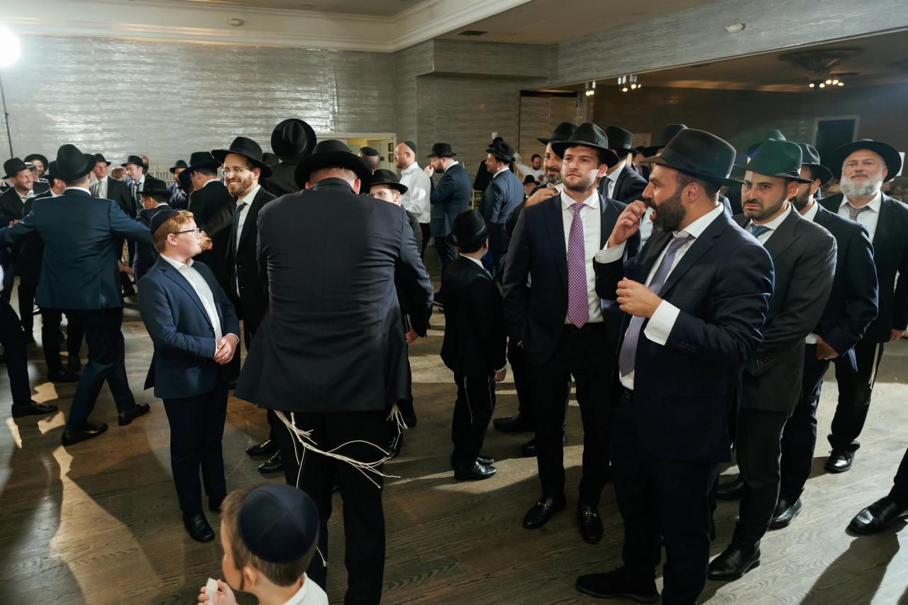 Guests dance in a crowded circle, photographed through a foreground of shoulders and hats. Bar Mitzvah photography at Eagle Ridge, Lakewood, NJ.