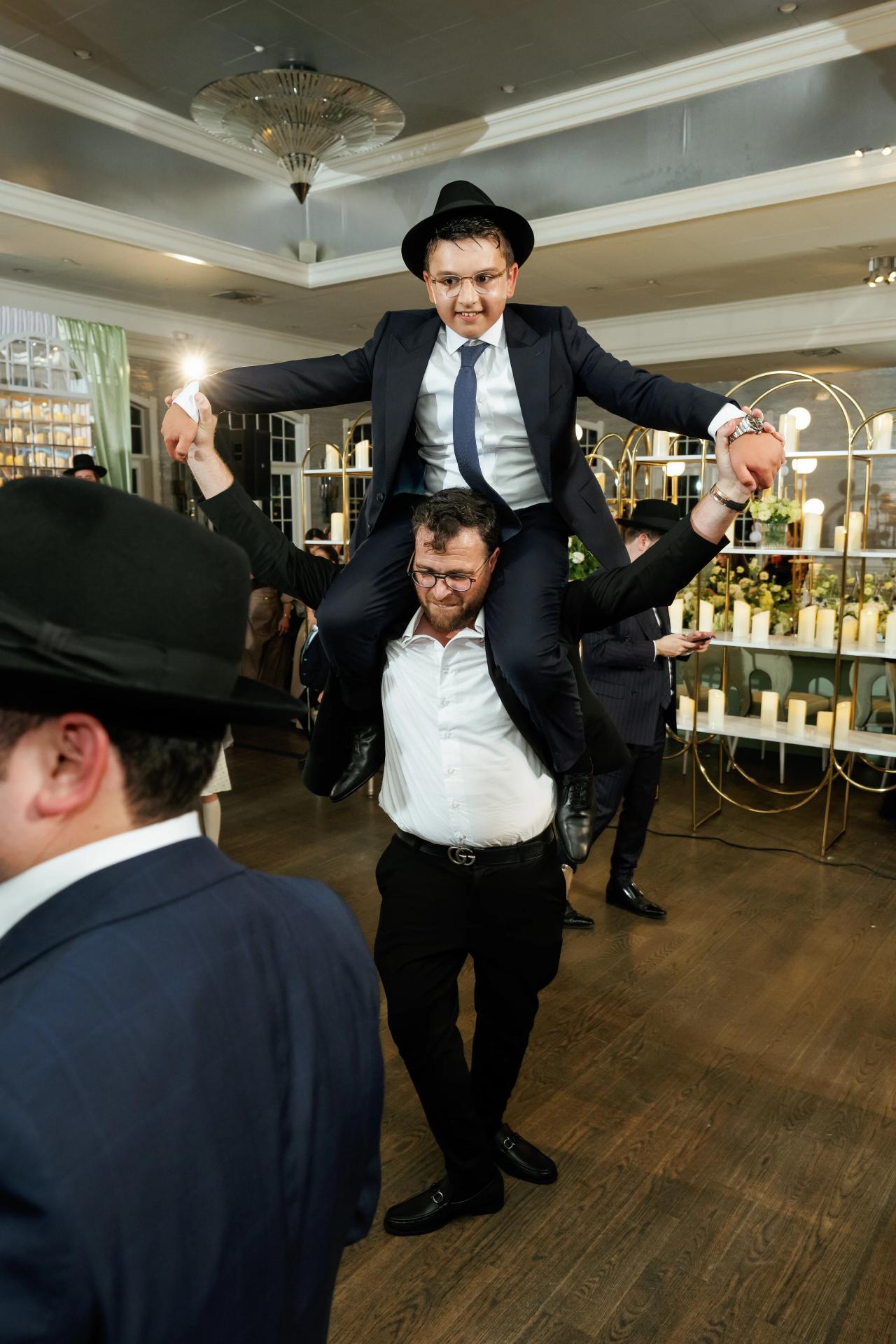 The bar mitzvah boy is carried on shoulders with his arms outstretched above the guests. Bar Mitzvah photography at Eagle Ridge, Lakewood, NJ.