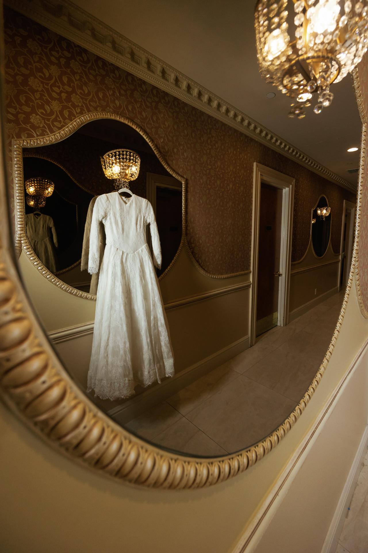 A wedding dress reflected in an ornate oval mirror during bridal preparation. Wedding photography at Ateres Charna, New Square, NY.