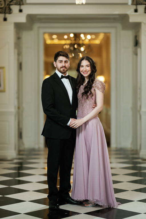 A formally dressed couple stands side by side in a lit ballroom corridor, smiling and looking at the camera.