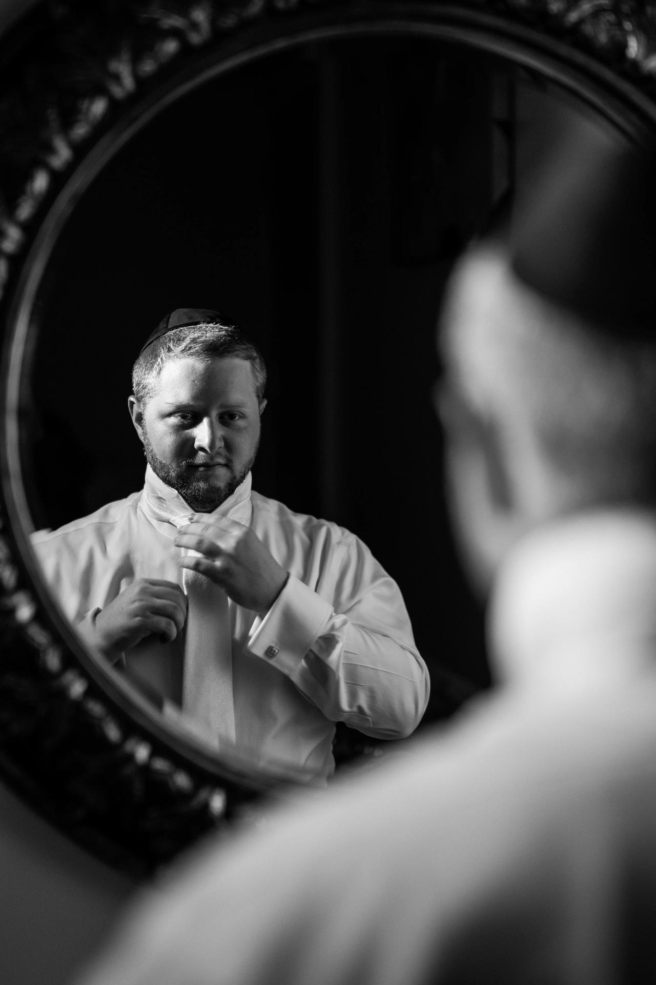 A black-and-white groom portrait reflected in an ornate mirror, with shoulders in the foreground. Wedding photography at Ateres Charna, New Square, NY.
