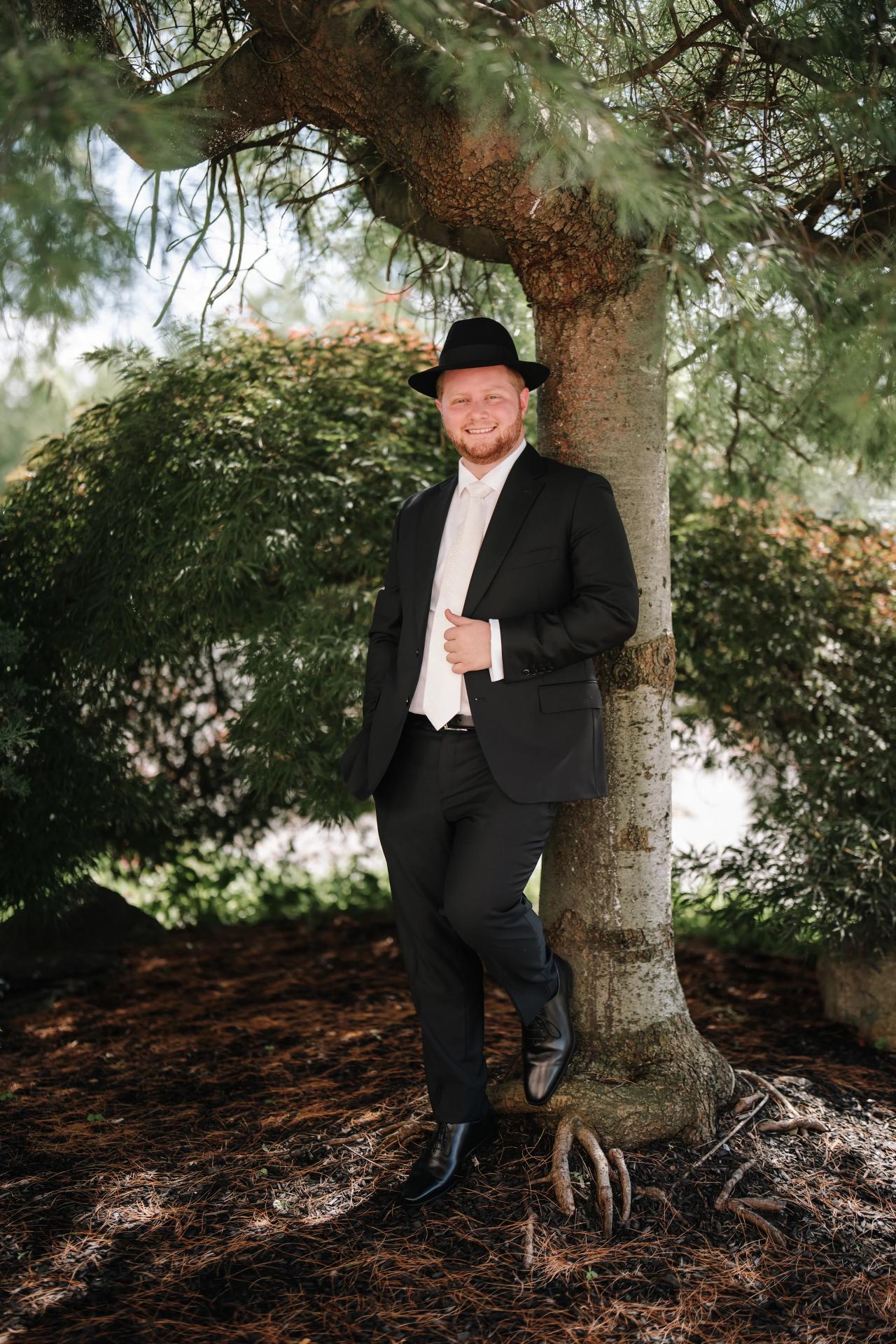 The groom standing beneath a tree, with garden greenery framing his formalwear. Wedding photography at Ateres Charna, New Square, NY.