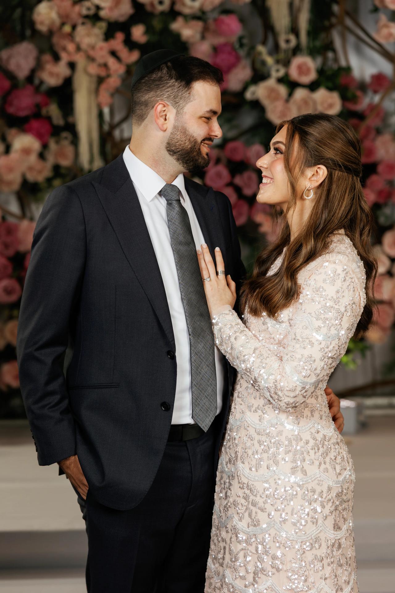 A candid family embrace and forehead kiss beside the wedding floral wall. Wedding photography at Ateres Charna, New Square, NY.