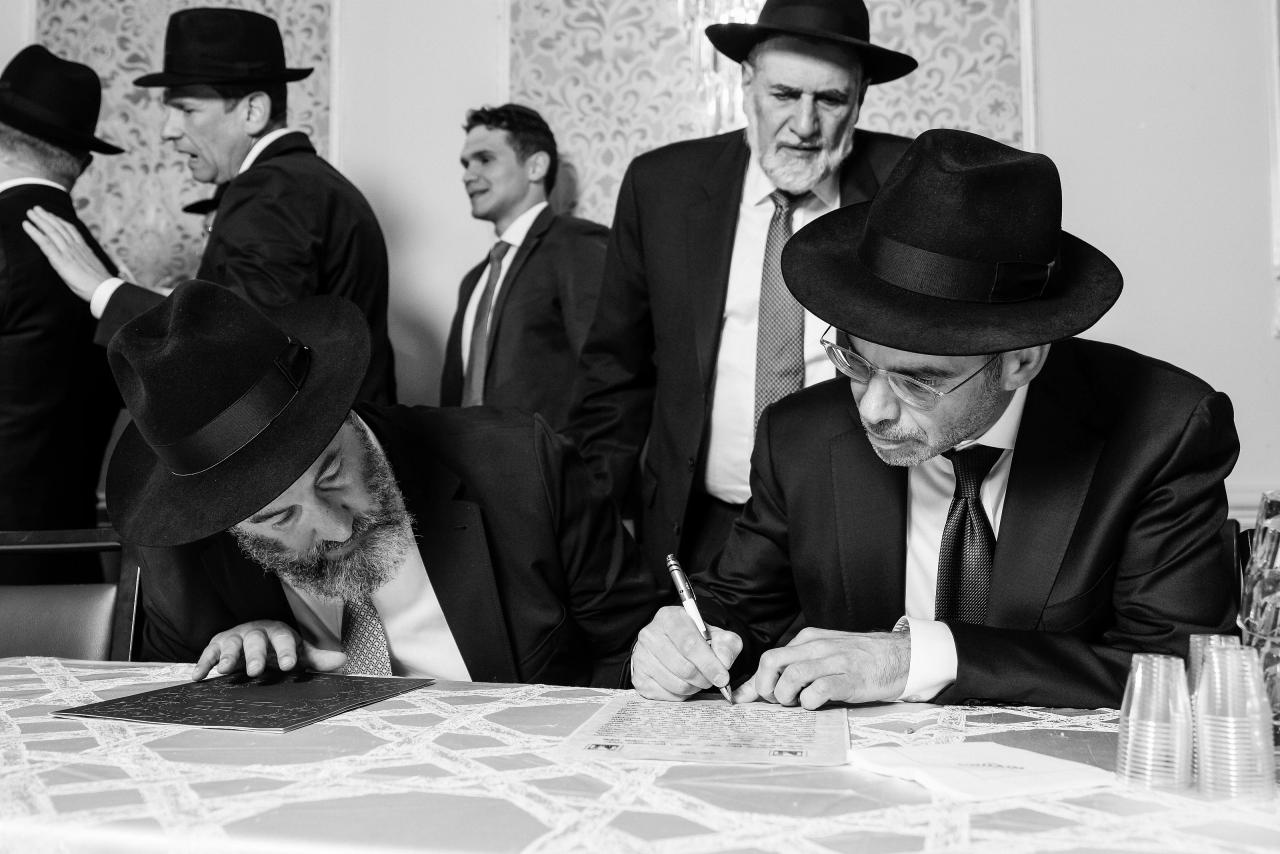 A black-and-white ketubah-signing scene with the groom intent on the page. Wedding photography at Ateres Charna, New Square, NY.