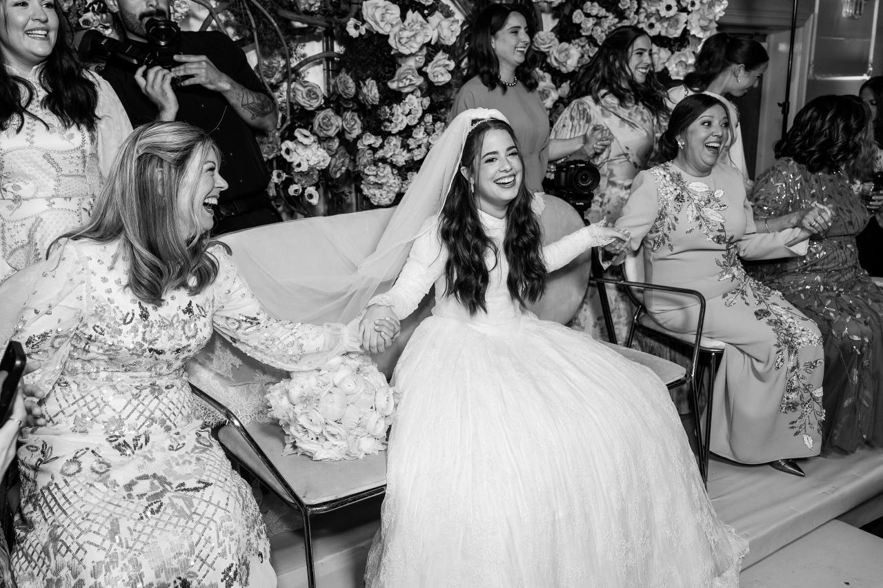 The bride smiling beneath her veil with arms extended, surrounded by friends in a black-and-white bedeken photograph. Wedding photography at Ateres Charna, New Square, NY.