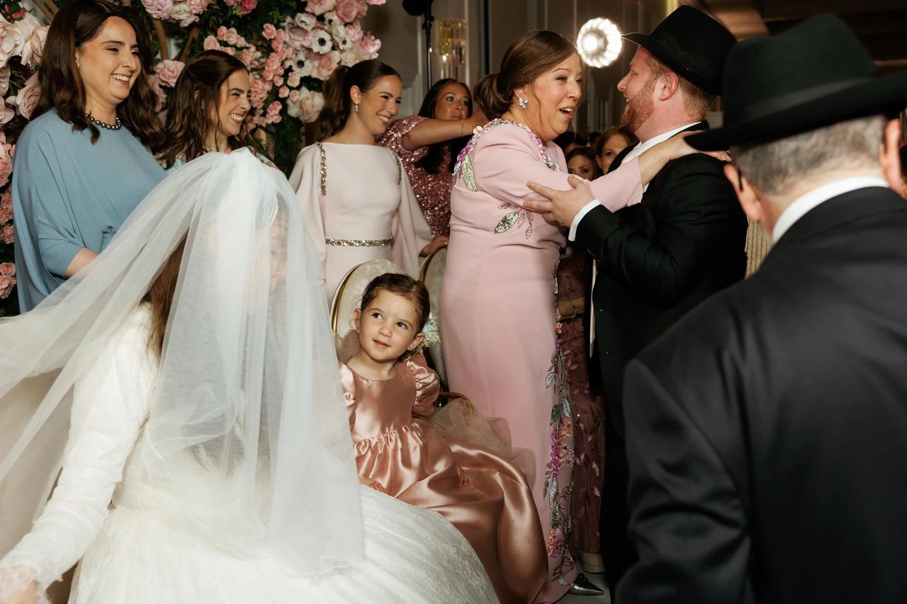 A family forehead kiss during the bedeken, with flowers and guests layered behind the bride. Wedding photography at Ateres Charna, New Square, NY.