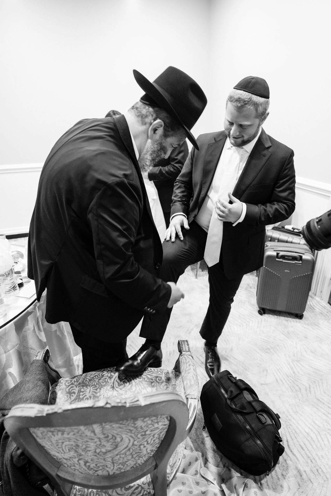 A close black-and-white view of the groom being helped into his ceremonial kittel. Wedding photography at Ateres Charna, New Square, NY.