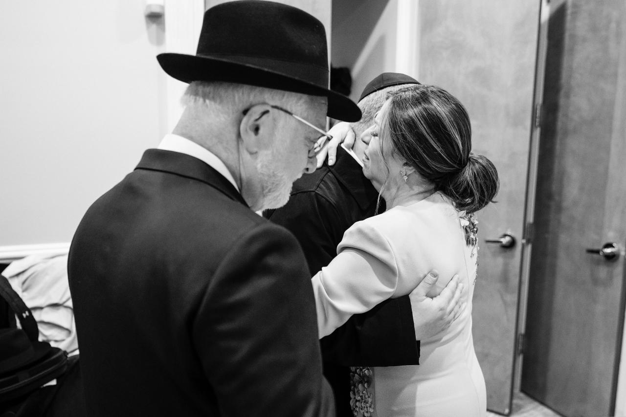 A quiet black-and-white family blessing, with faces close and hands holding the groom. Wedding photography at Ateres Charna, New Square, NY.