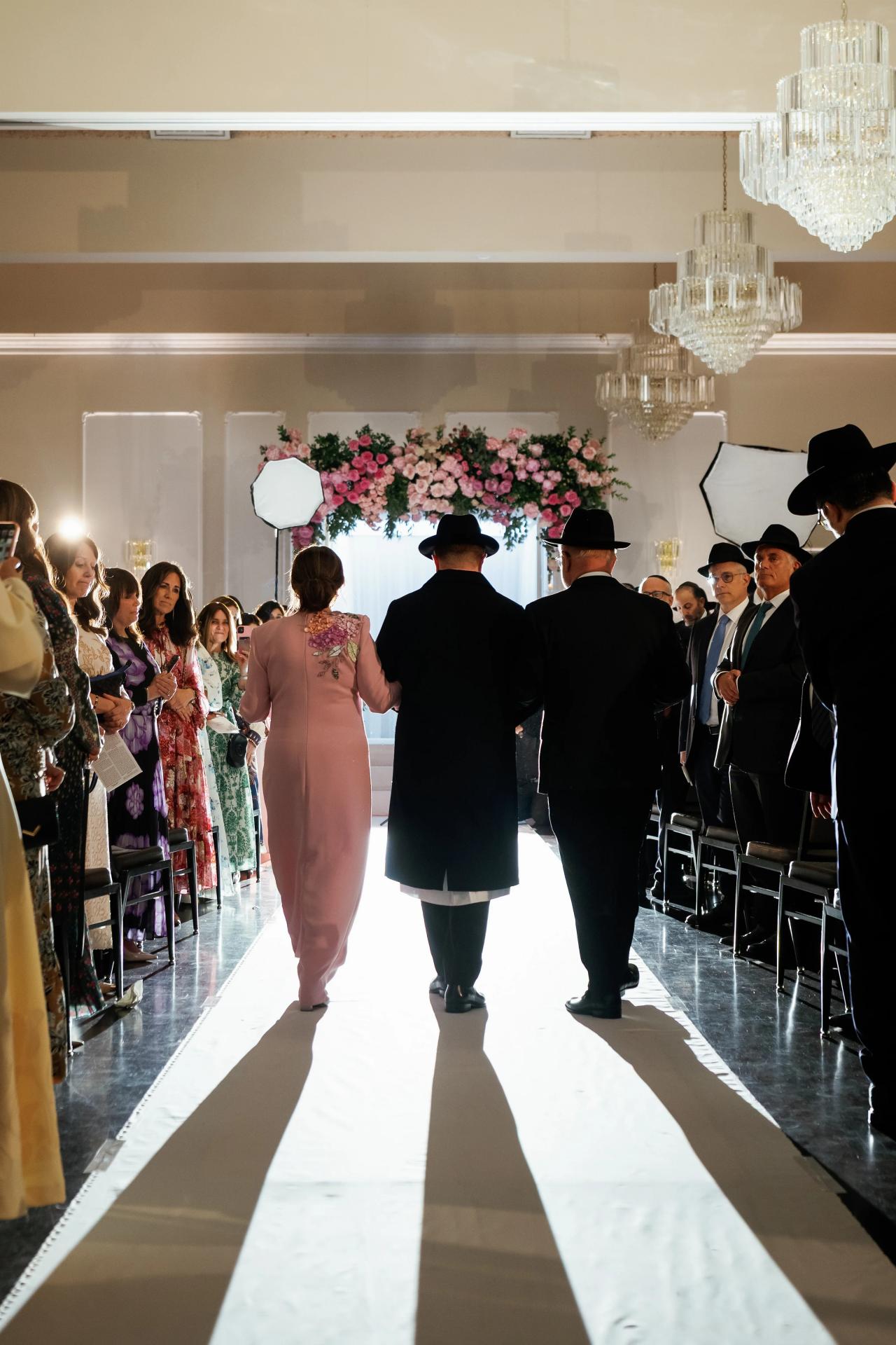 A wedding processional along a sunlit indoor aisle, with guests silhouetted at the edges. Wedding photography at Ateres Charna, New Square, NY.
