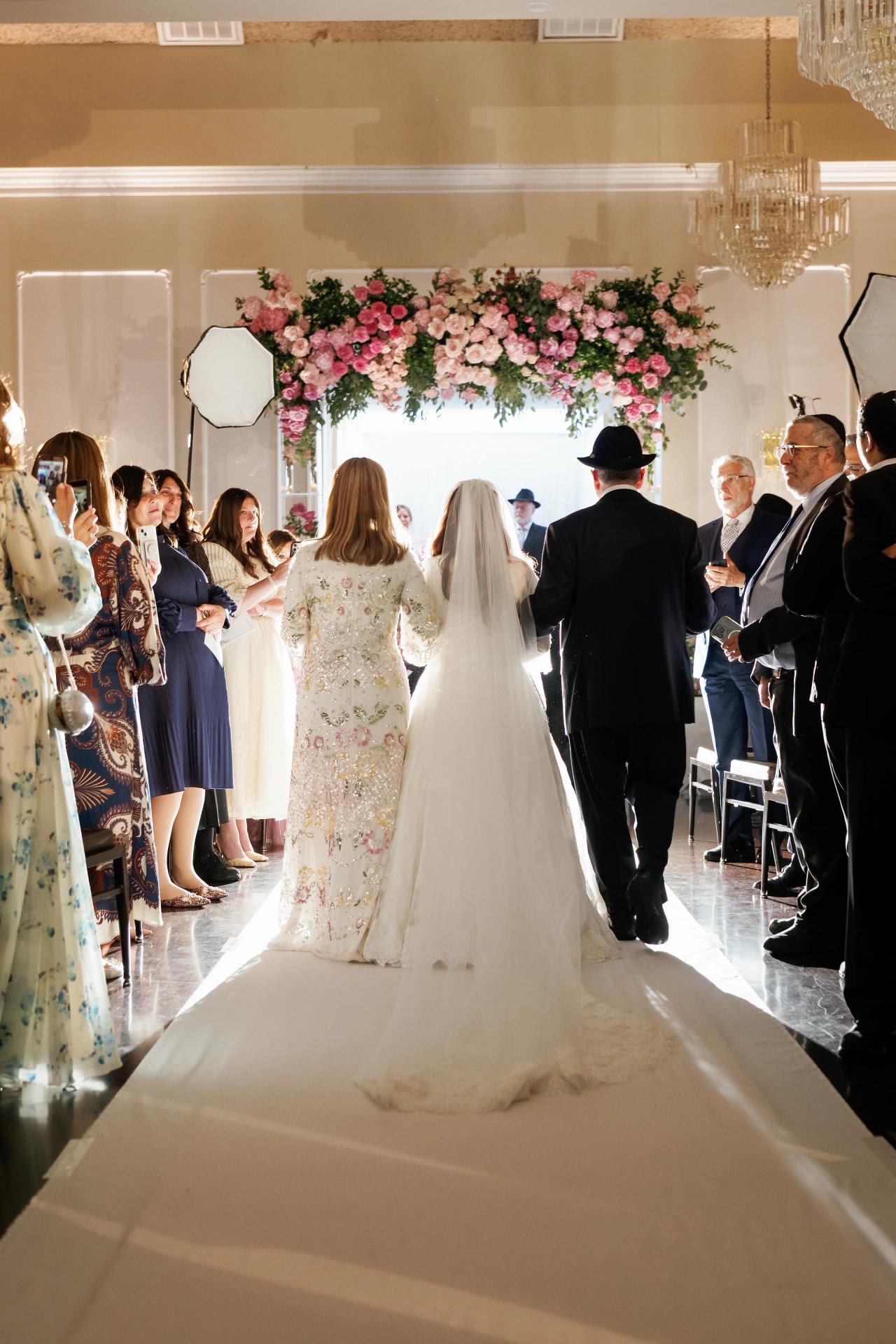 The bride entering beneath a floral canopy, her long veil catching the window light. Wedding photography at Ateres Charna, New Square, NY.