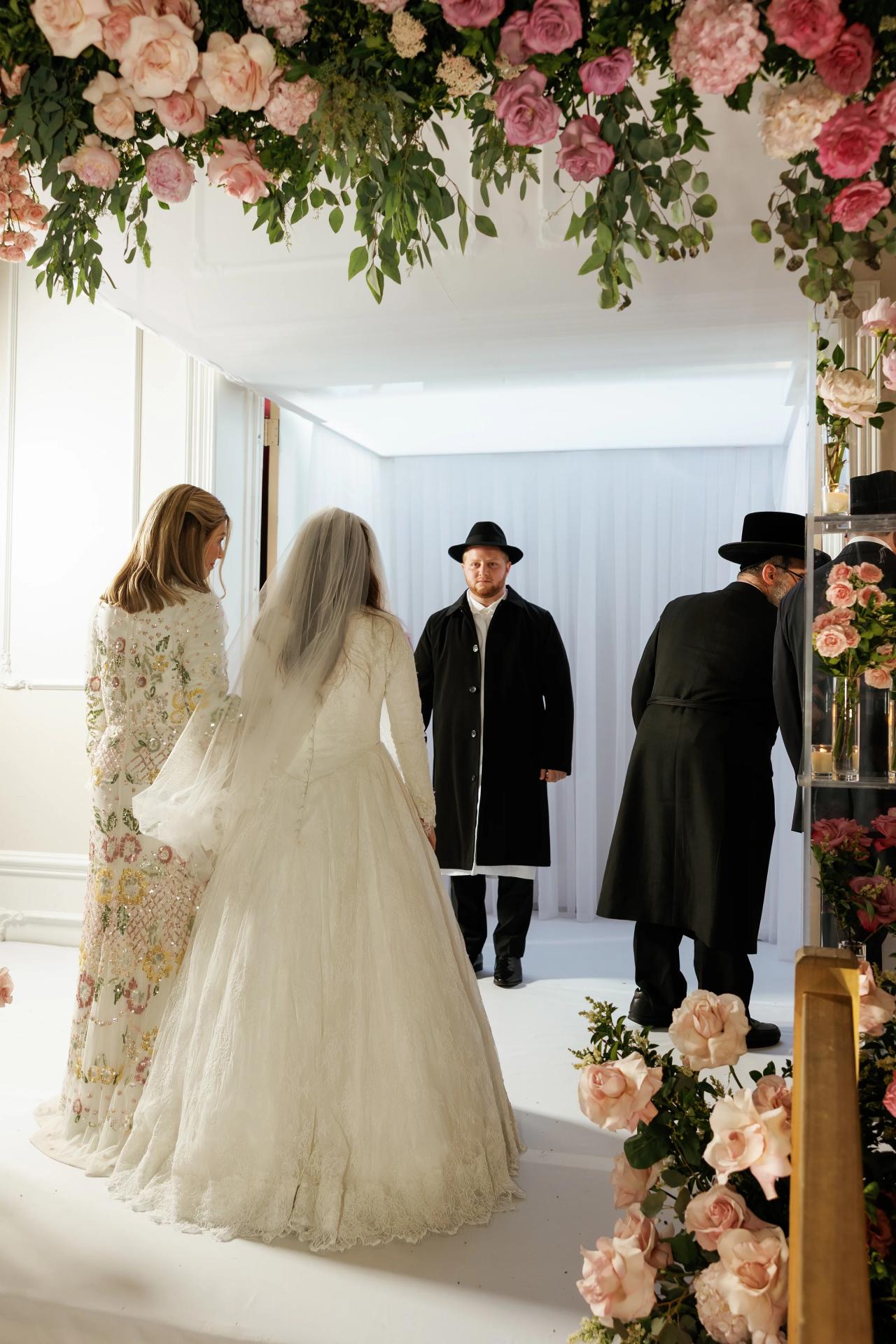 The bride approaching the indoor chuppah, seen from behind against luminous white curtains. Wedding photography at Ateres Charna, New Square, NY.
