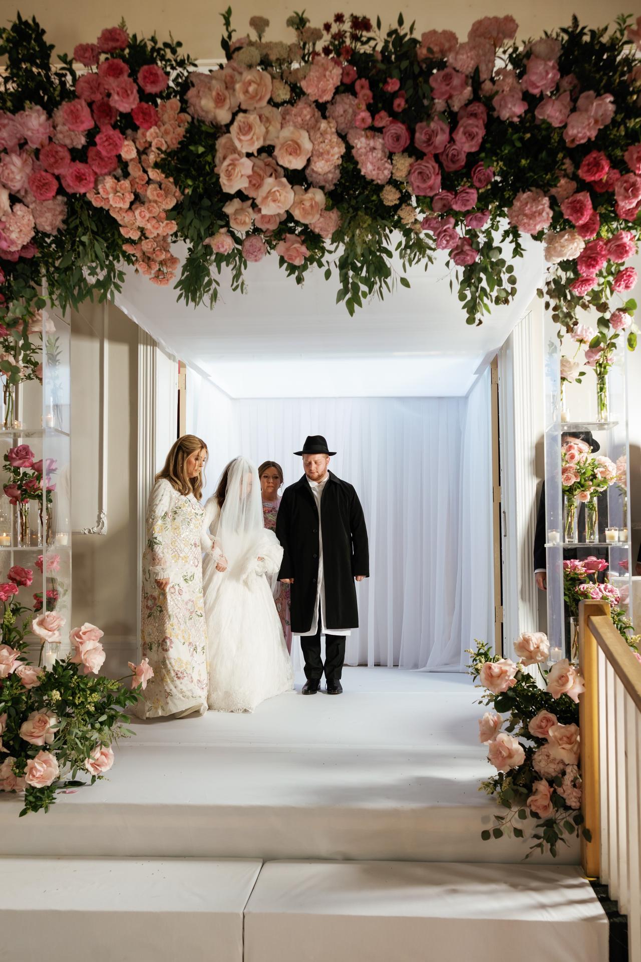 A wide symmetrical view of the indoor floral chuppah against luminous white curtains at Ateres Charna. Wedding photography.