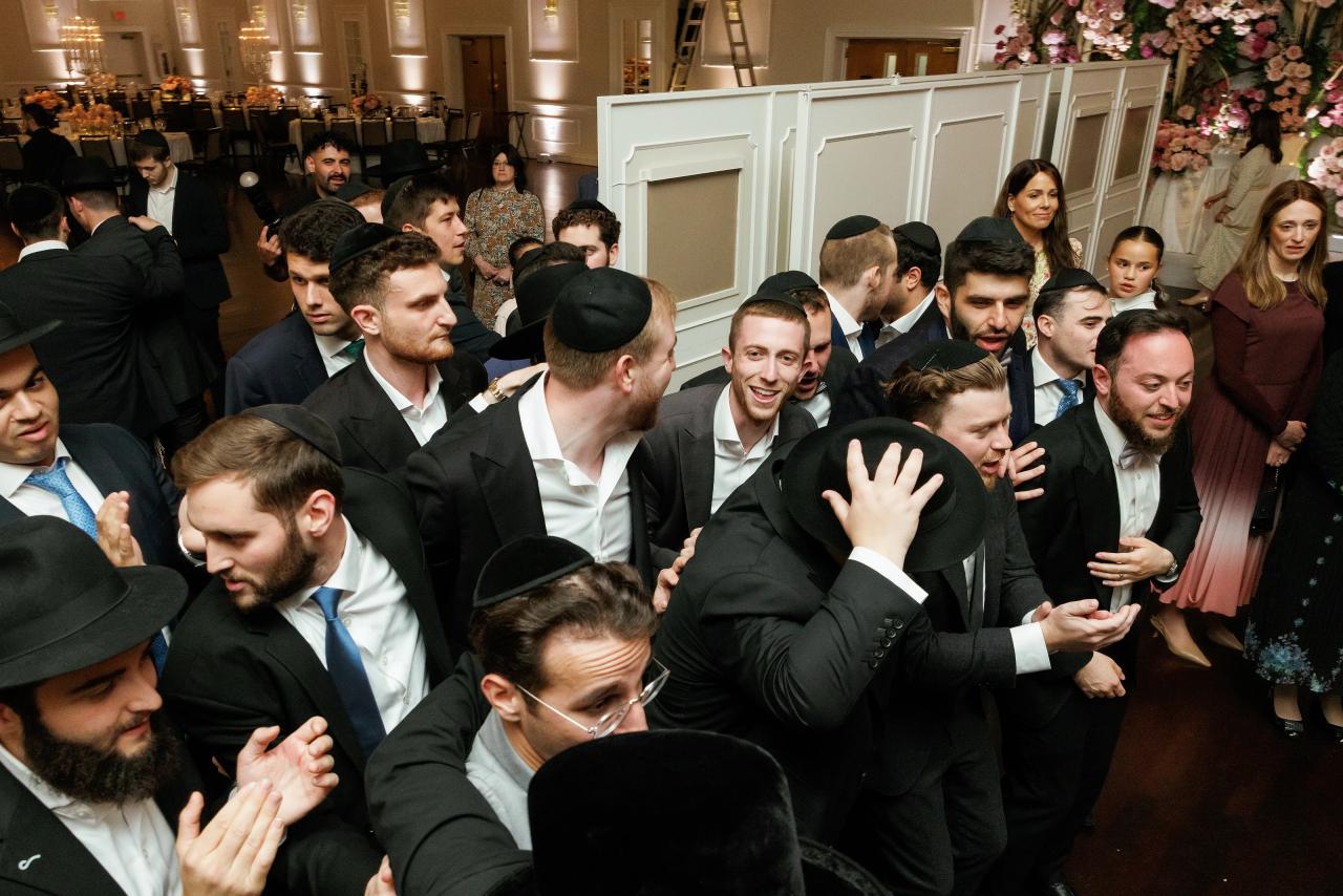 The groom surrounded by cheering guests in a lively black-and-white wedding photograph. Wedding photography at Ateres Charna, New Square, NY.