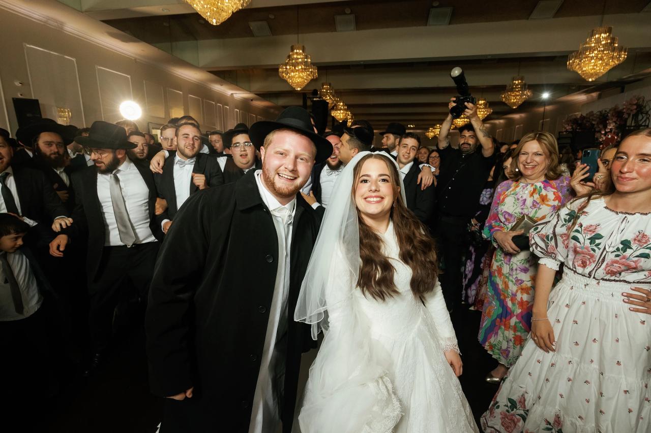 The smiling bride and groom walking through a crowd of celebrating guests at Ateres Charna, with raised hands and chandeliers behind. Wedding photography.